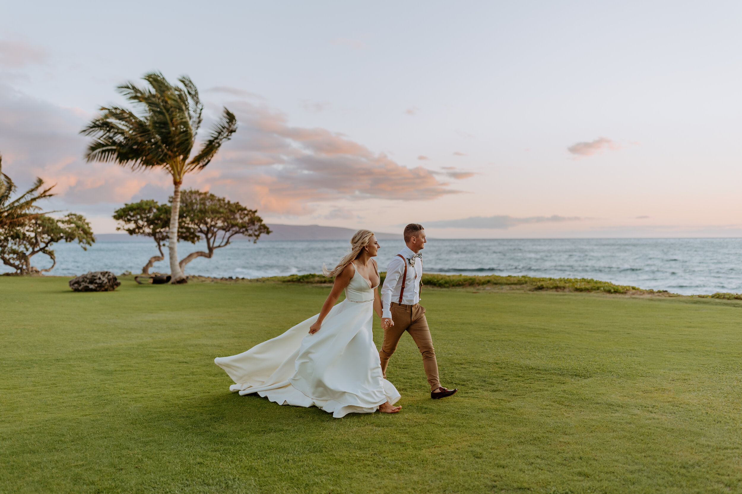 maui bride and groom wedding day walking on a beach with palm trees and the sunset ocean behind them