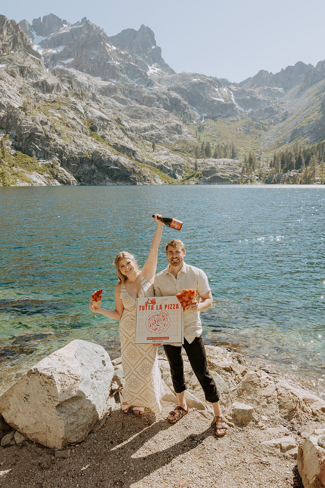 Couple holding hands while hiking to an alpine lake in the Eastern Sierras Custom pizza box engagement picnic with a mountain backdrop Candid engagement photo of couple sharing pizza by a lake Couple popping champagne next to friends and paddle boards at a secret lake near Tahoe Dog snuggled between engaged couple on a lakeside rock Friends celebrating engagement with floaties and paddle boards on alpine lake Couple laughing in the water fully clothed after jumping in post-engagement shoot Mountain engagement photo at golden hour in the Sierra Nevada range Playful engagement moment with dog, pizza, and champagne in the California mountains