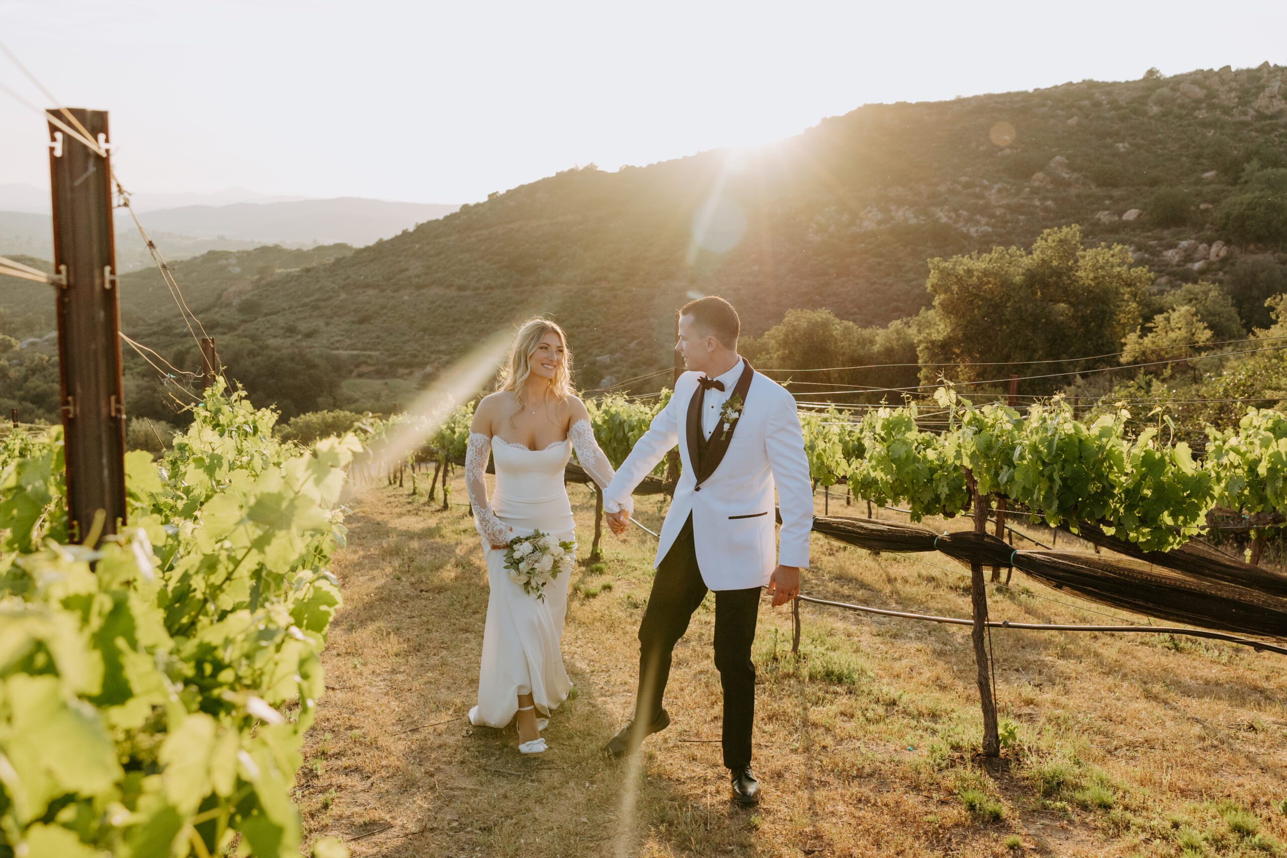 Bride arriving to ceremony in vintage green convertible at Milagro Winery Romantic ceremony under oak trees at Milagro Winery in Ramona, CA Bride and groom sharing vows under shaded SoCal oaks Couple walking through vineyard at sunset with golden hills in the background San Diego wedding photographer capturing candid winery moments Reception outside the Crush House under market lights Cocktail hour inside stone wine cave at Milagro Winery Cypress tree-lined dirt road portraits at Milagro Bride in modern silhouette dress holding hands with groom Guests dancing under string lights at Milagro Winery reception Gelato cart and bar trailer at winery wedding in California Bride and groom riding golf cart through winery for golden hour photos