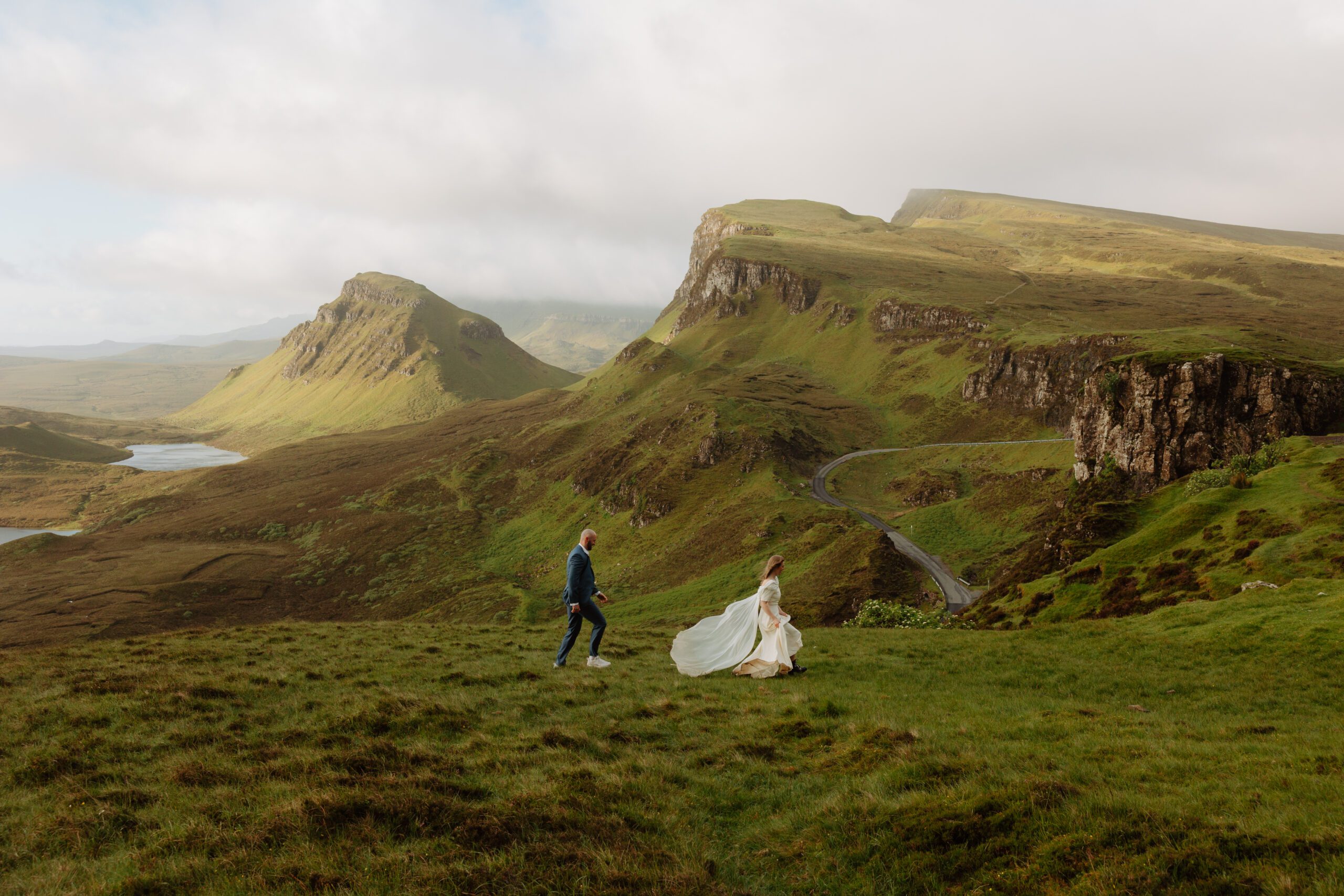 Couple standing on a cliff at sunrise on the Isle of Skye, wrapped in soft golden light and Highland mist Romantic anniversary photo session in the Scottish Highlands with dramatic mountain views Elopement-style shoot on the Isle of Skye with moody skies and a glowing sunrise Scottish couple embracing on a windy mountain with the ocean in the distance Bride in a flowy white dress walking through mossy grasslands at dawn Couple laughing together in front of rugged Highland cliffs during a destination photo session Golden hour light breaking through misty mountains during a romantic couple’s session in Scotland Intimate anniversary shoot in the Highlands with soft, ethereal lighting and a scenic view